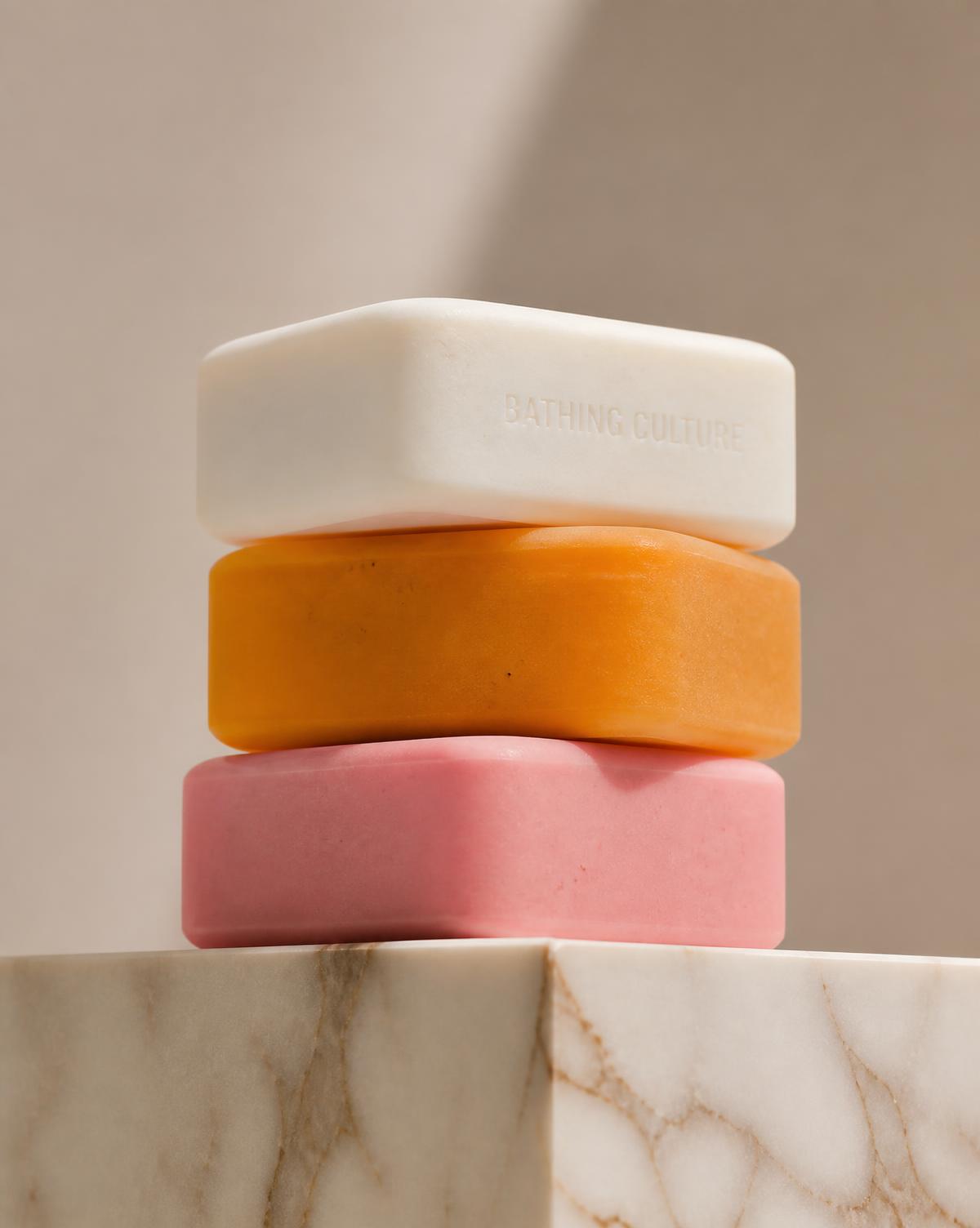 Three Lumière Bar soaps in white, orange, and pink stacked on a marble plinth